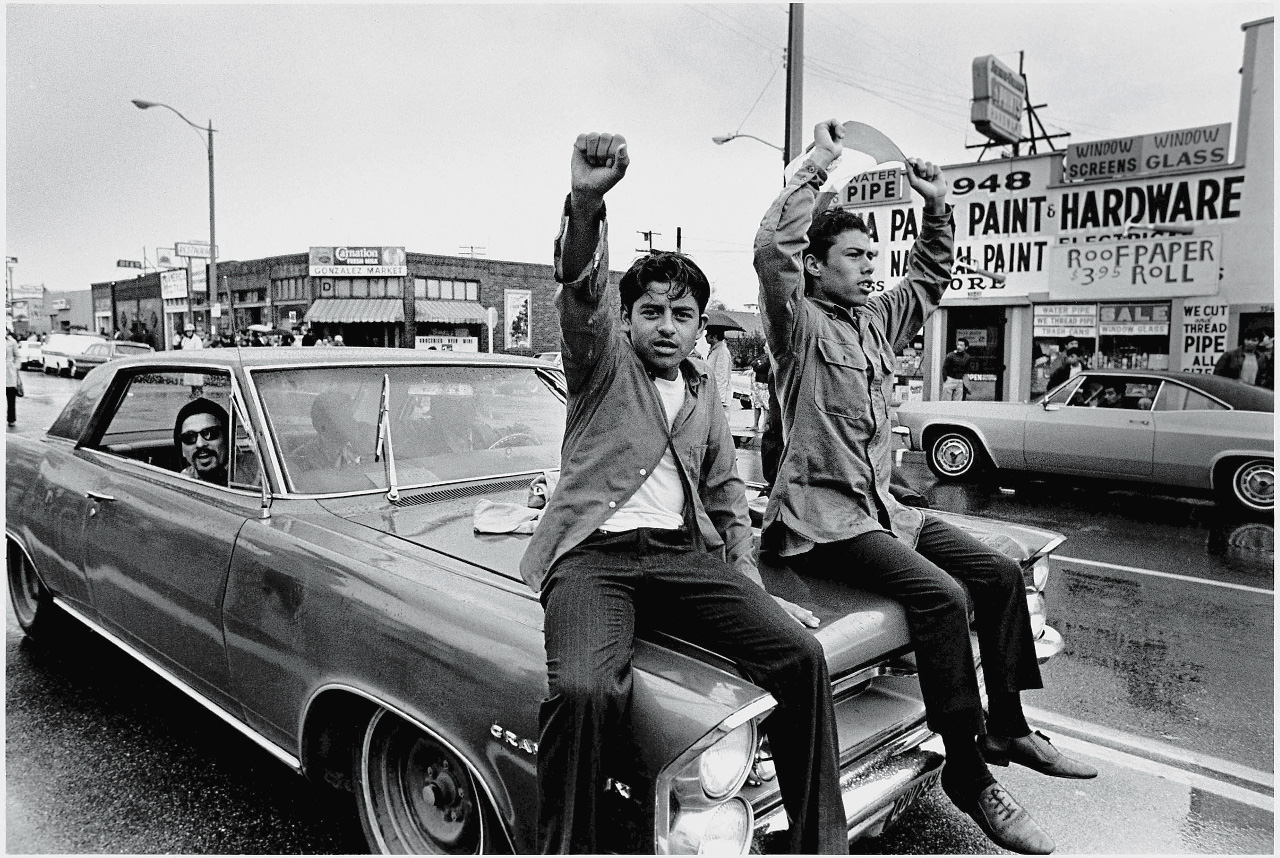 Two young Chicano men ride on the hood of a car and raise their fist during a National Chicano Moratorium Committee march in opposition to the war in Vietnam, Los Angeles, California, February 28, 1970. (Photo by David Fenton/Getty Images)